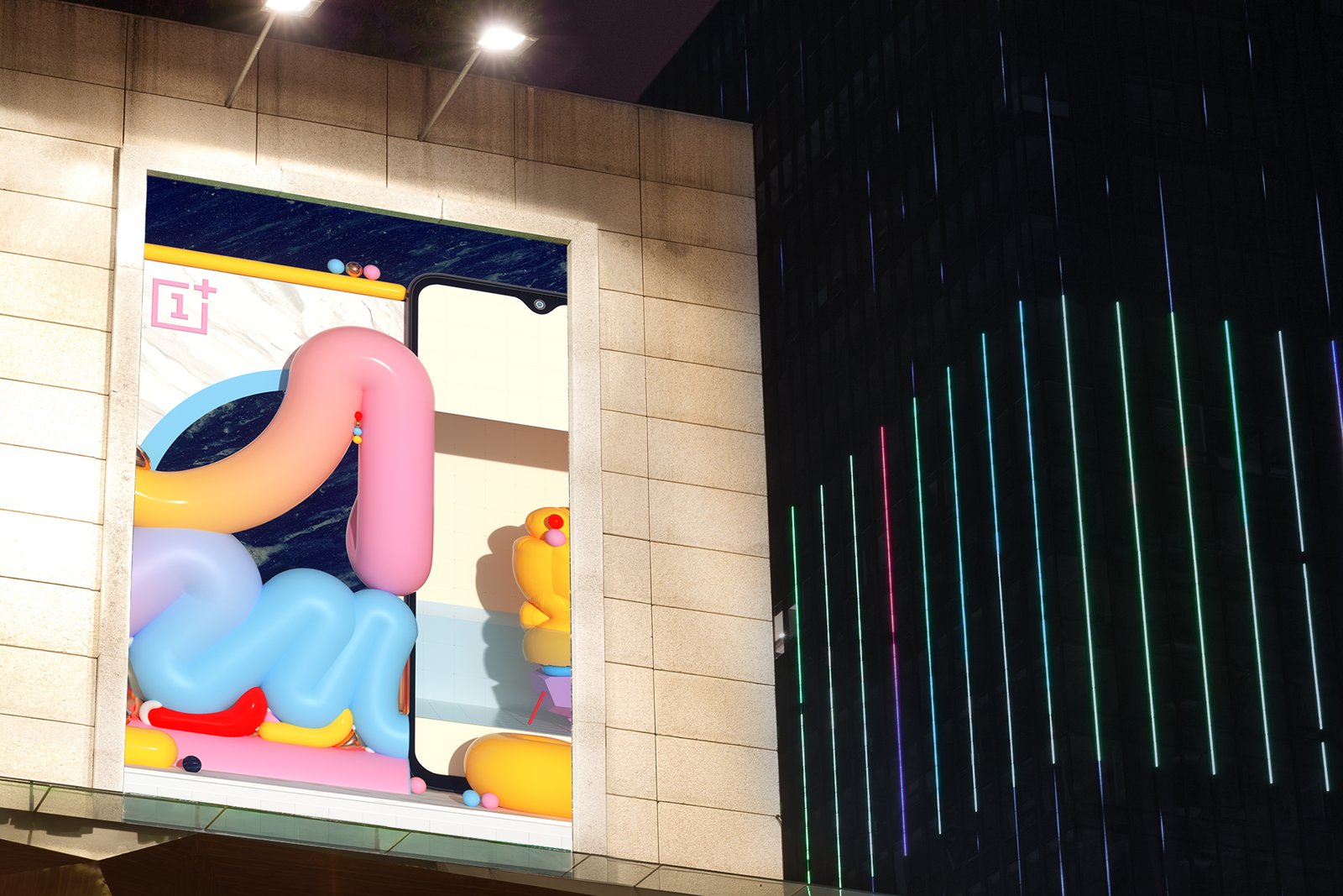 building-side billboard at night: pastel inflatable shapes wrapping a oneplus phone